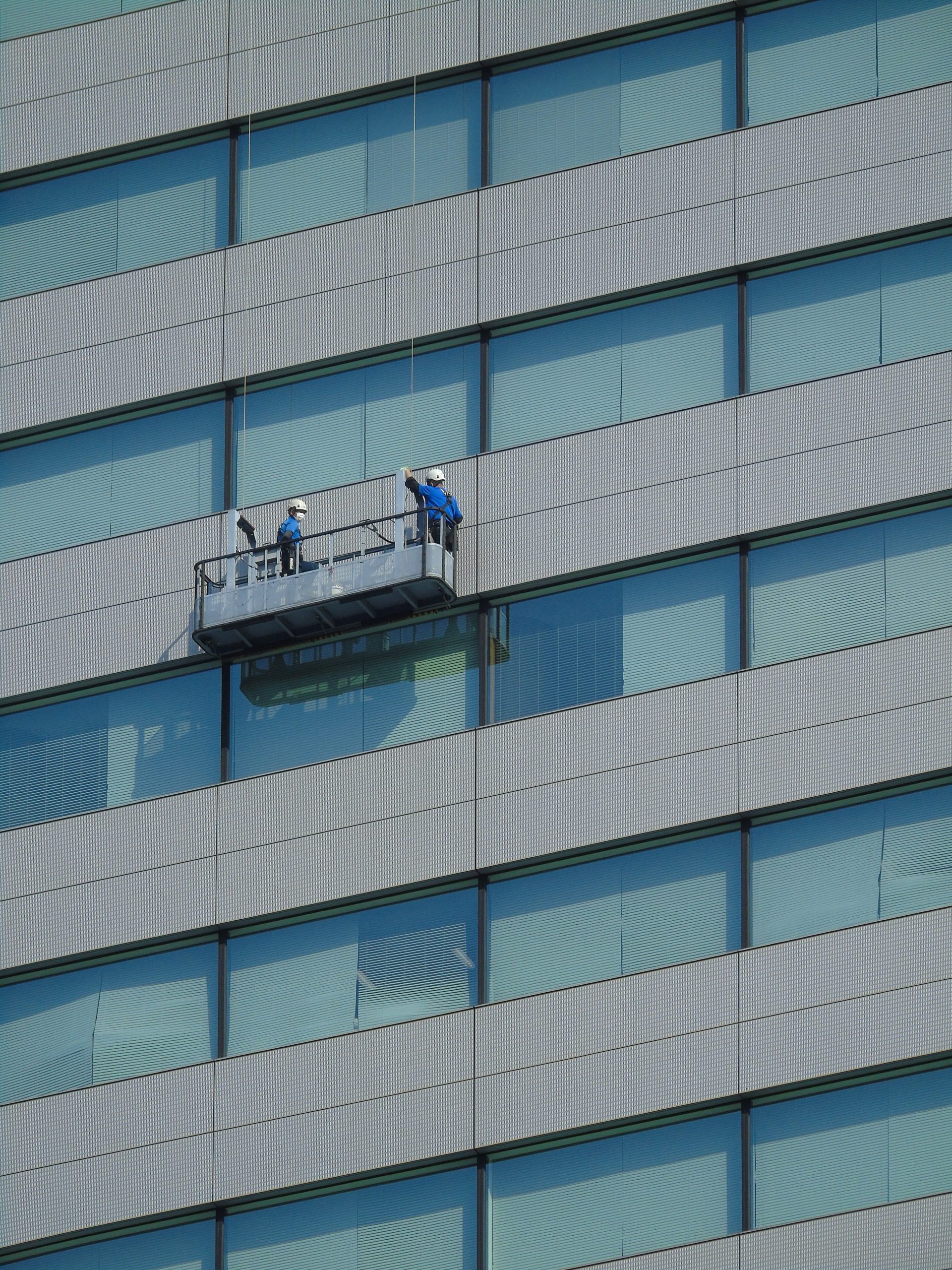 Zwei Arbeiter reinigen die Fenster einer modernen Glasfassade von einer Hängeplattform aus.