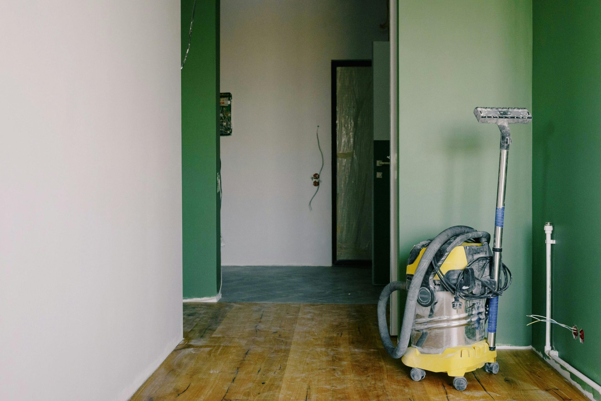 Industriestaubsauger auf schmutzigem Parkett in einem Zimmer mit grünen Wänden und Türöffnung in einer geräumigen Wohnung während Renovierungsarbeiten